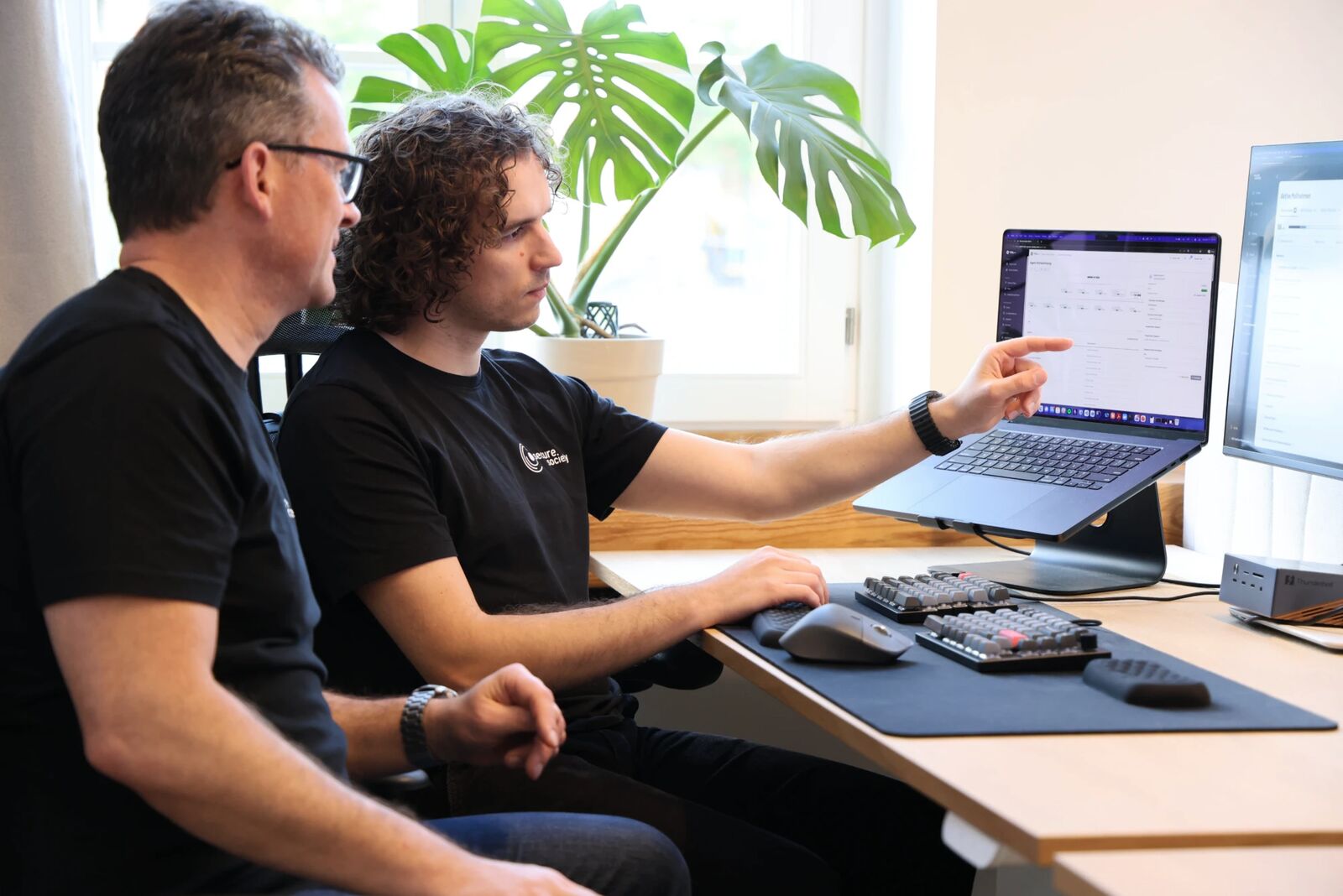 Zwei Personen sitzen am Schreibtisch und zeigen konzentriert auf die geöffnete Plattform auf dem Laptop