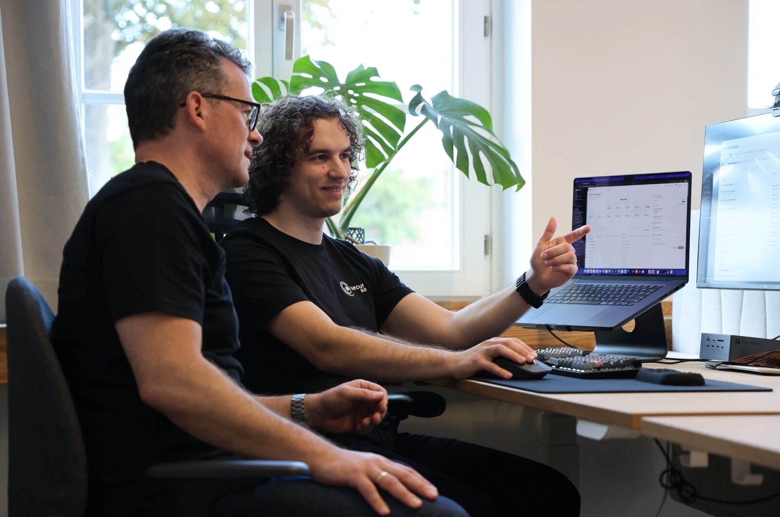 Zwei Personen am Schreibtisch am Fenster, eine lächelt und zeigt auf die Plattform auf dem Laptop